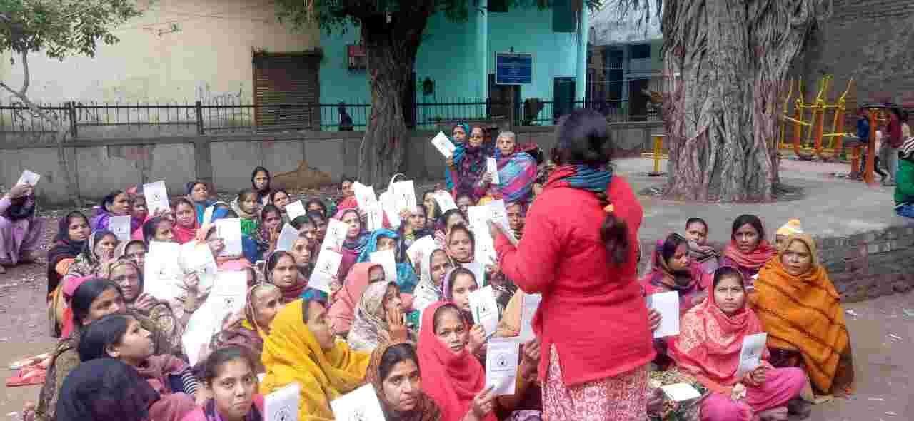 A group of women seated outdoors holding documents while a facilitator speaks.
