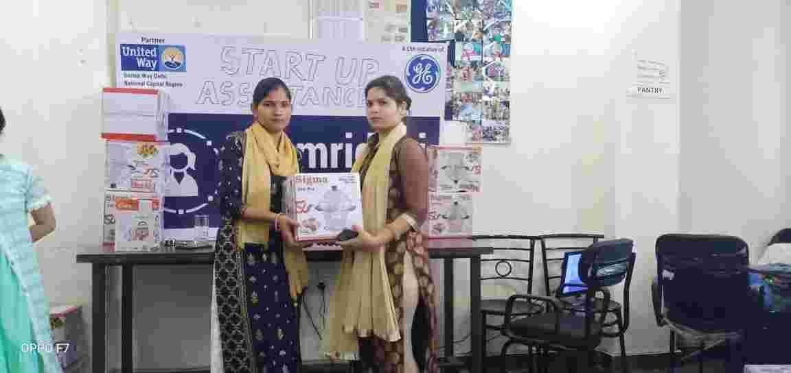 Two women standing together and holding a boxed item in front of a Samriddhi start-up assistance display.